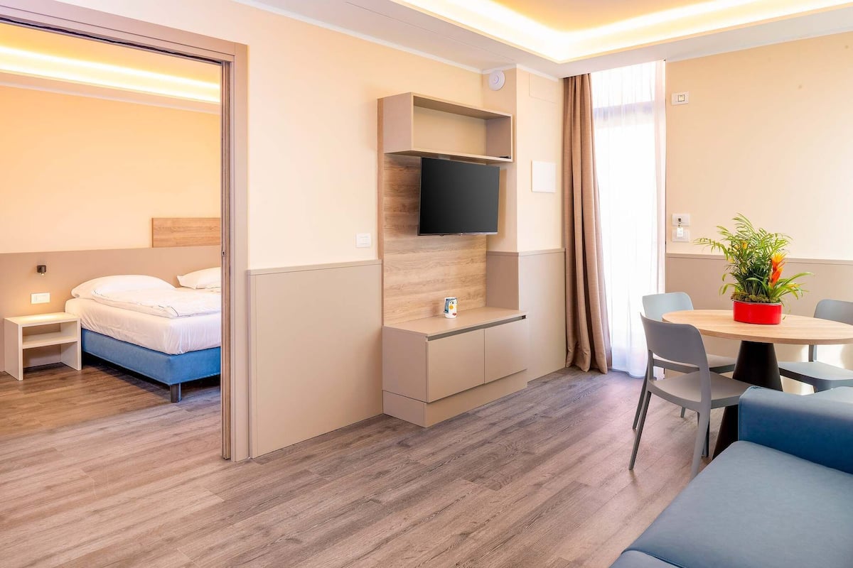 Une chambre d’hôtel moderne avec un lit, un téléviseur fixé au mur, une petite table avec des chaises et une plante en pot.