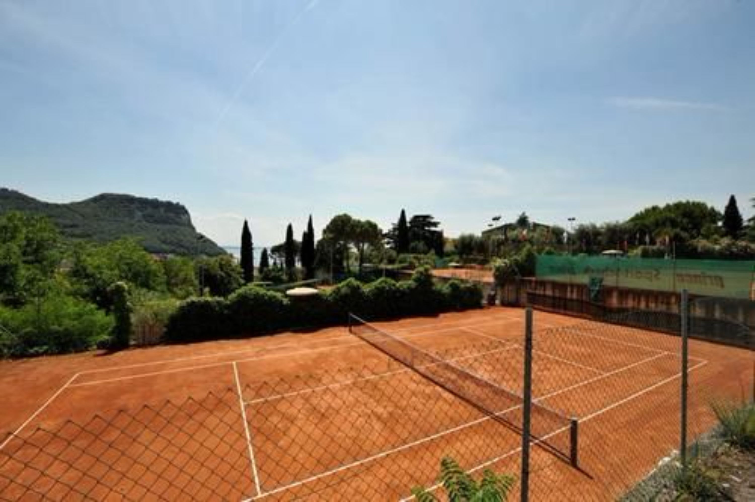 A tennis court with a fence, surrounded by greenery and hills.