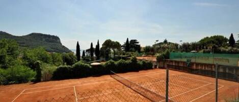 A tennis court with a fence, surrounded by greenery and hills.