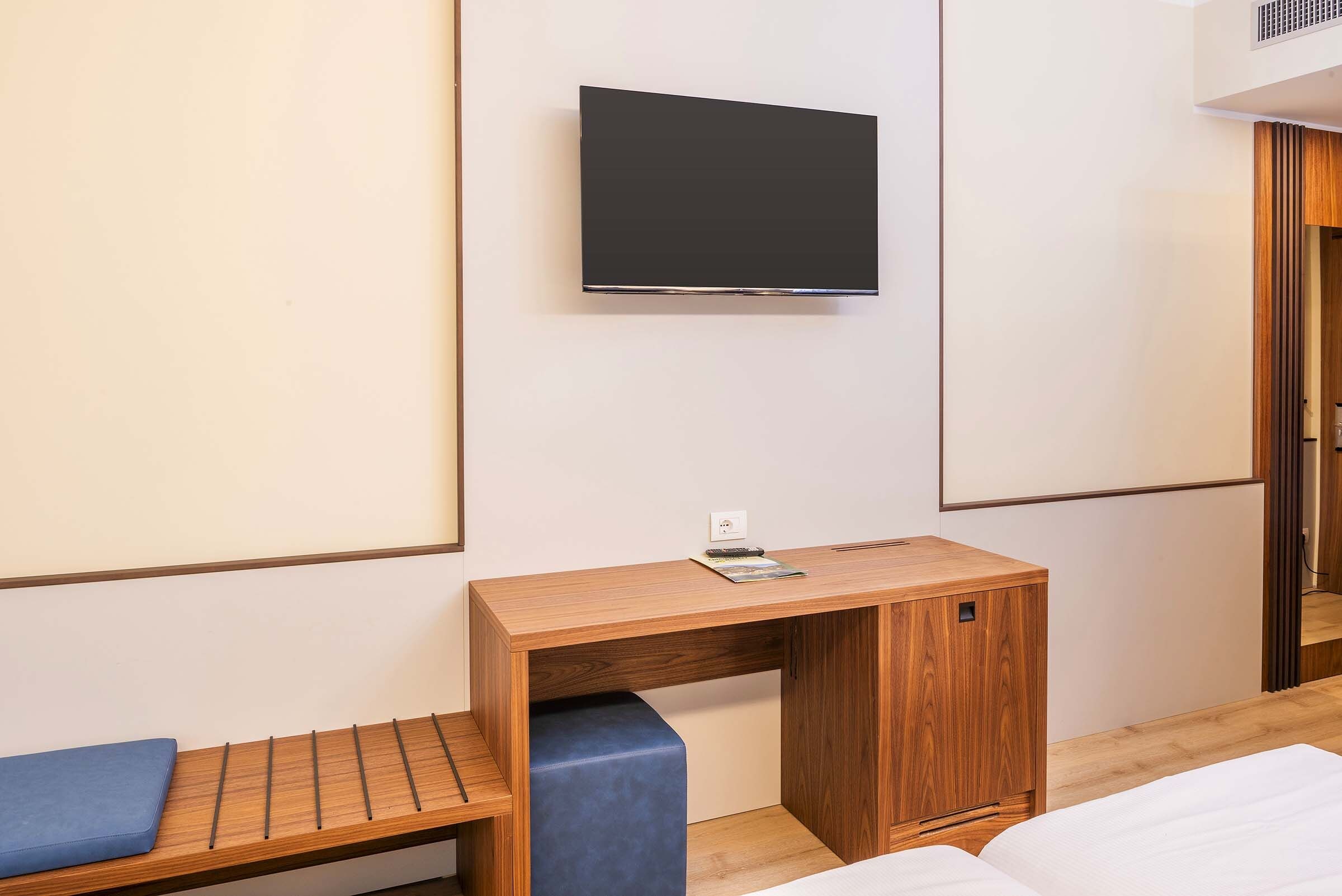 A hotel room with a wooden desk, a blue bench, a bed, and a mounted television.