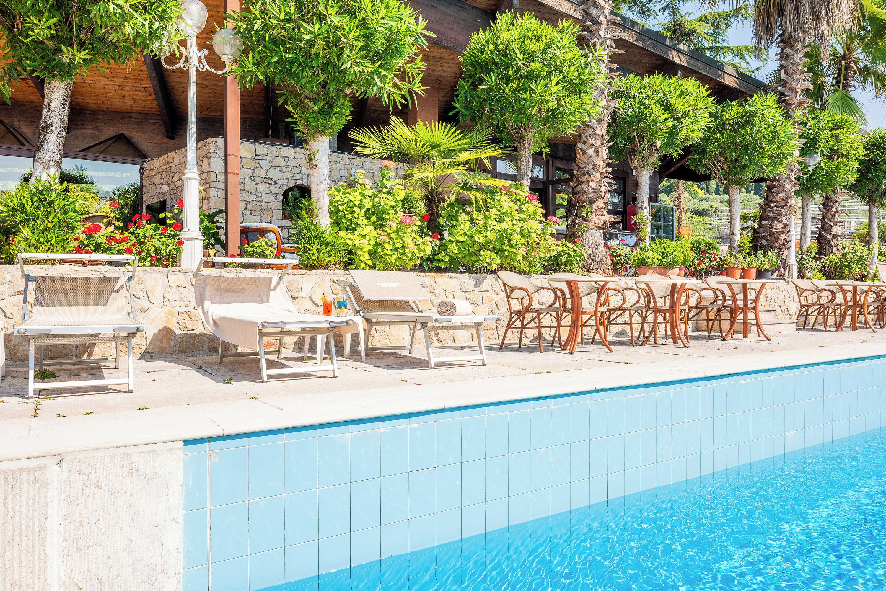 A poolside area with lounge chairs, a stone wall, and a variety of plants and trees.