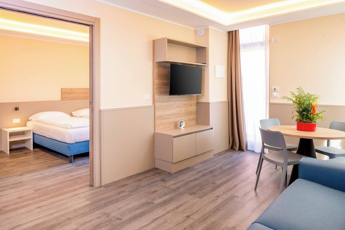 Une chambre d’hôtel moderne, équipée d’un lit, d’un téléviseur fixé au mur, d’une petite table avec des chaises et d’une plante en pot.
