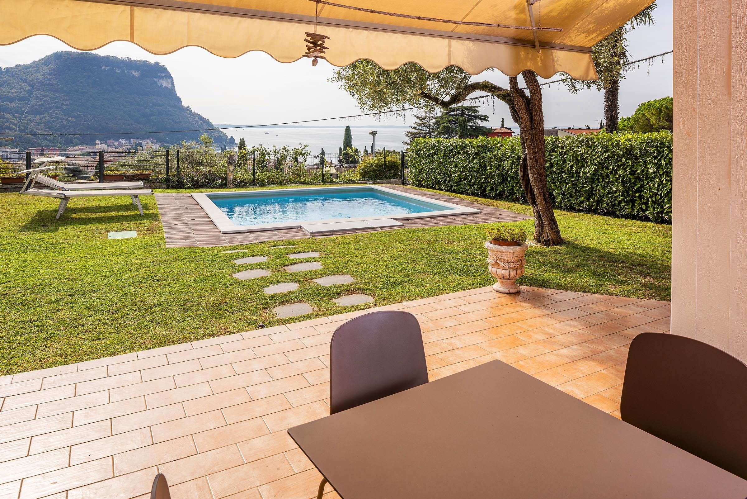 Ein Poolbereich mit Blick auf einen Berg und das Meer, eine überdachte Terrasse mit Tisch und Stühlen sowie ein gepflegter Rasen.