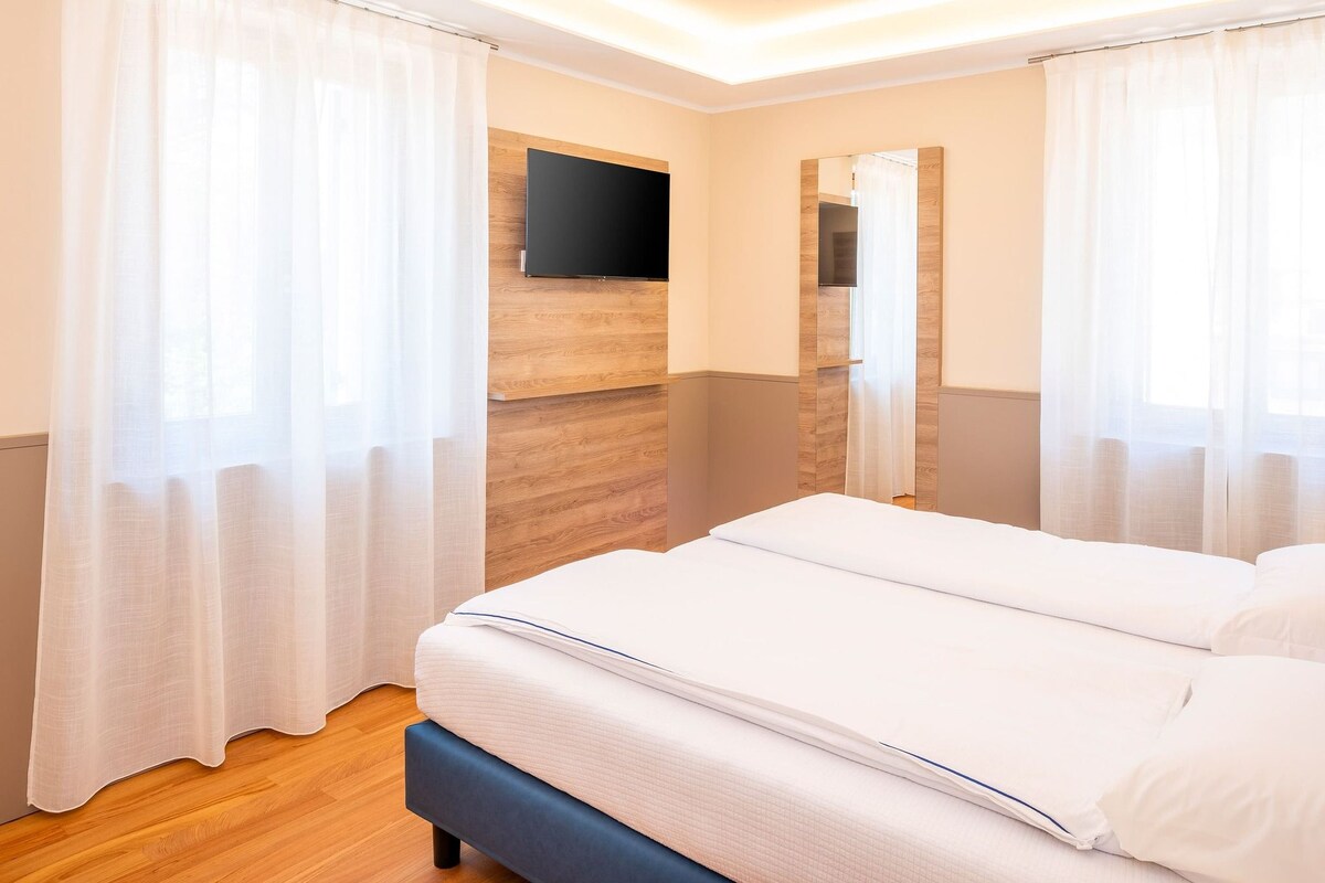 A hotel room with a large bed, wooden paneling, a flat-screen TV, and sheer curtains.