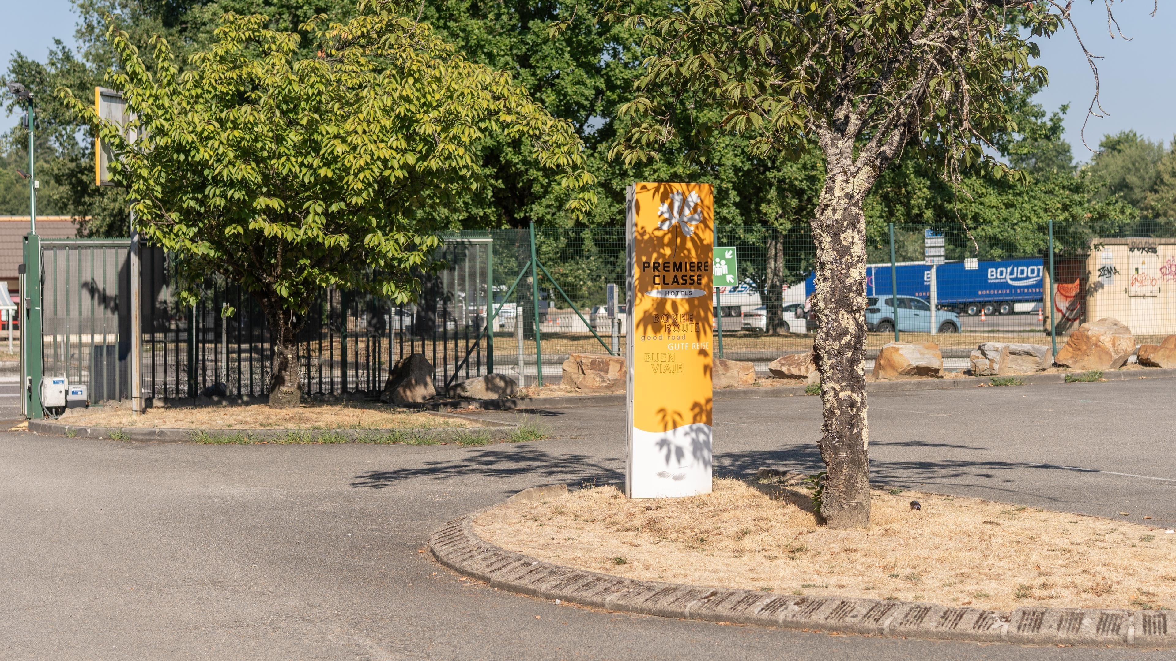 Un cartel con texto en varios idiomas, un árbol, una valla y una carretera.