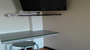 A small office space with a glass desk, a stool, a mounted flat-screen TV, and a towel on a shelf. - Premiere Classe Limoges Nord (Limoges)