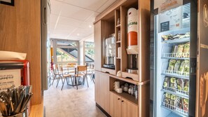 A café with a coffee machine, seating area, and a refrigerator stocked with snacks. - Premiere Classe Limoges Nord (Limoges)