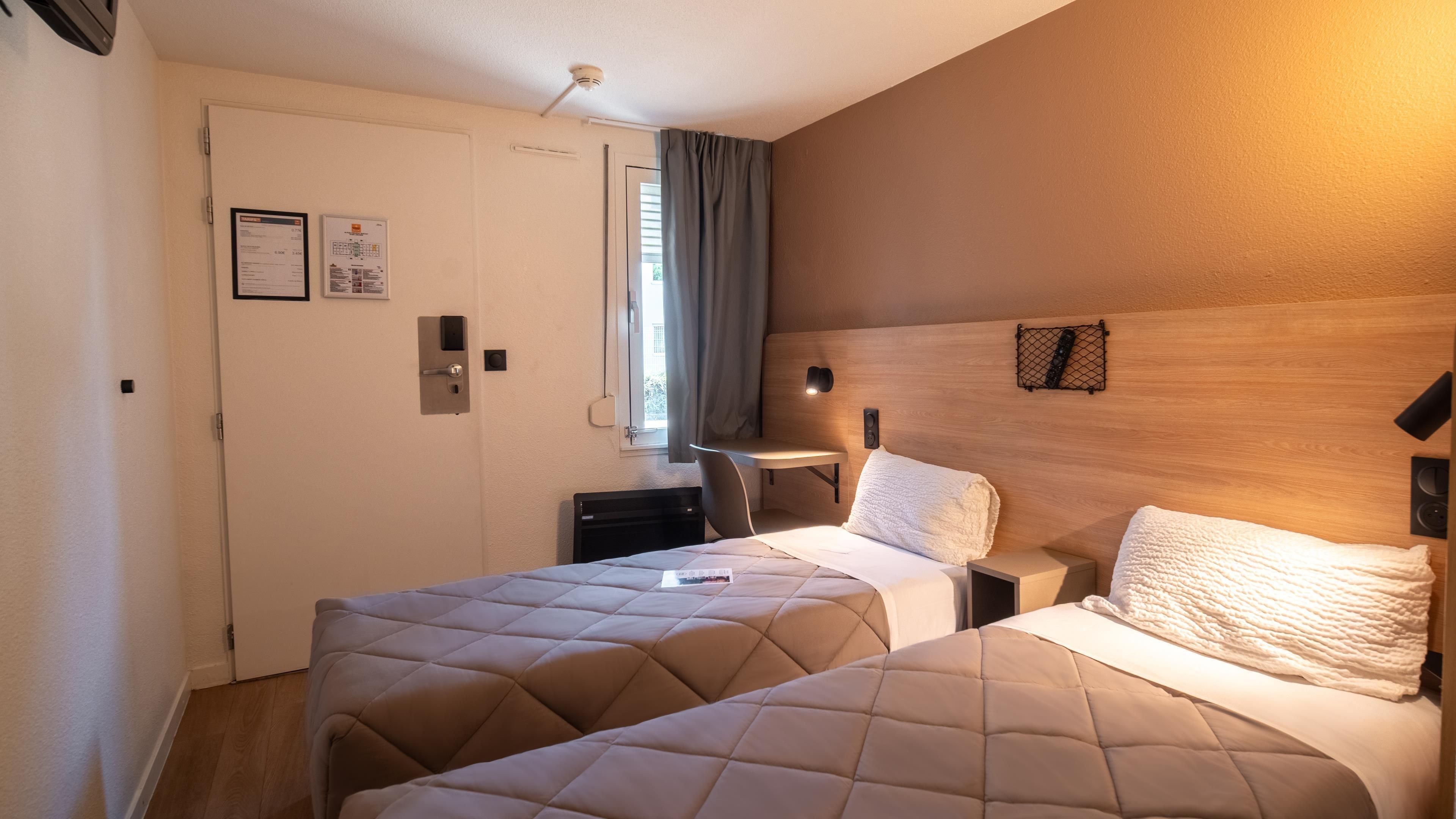 Ett hotellrum med två sängar, ett skrivbord, en stol, ett fönster med gardiner och en väggmonterad lampa.
