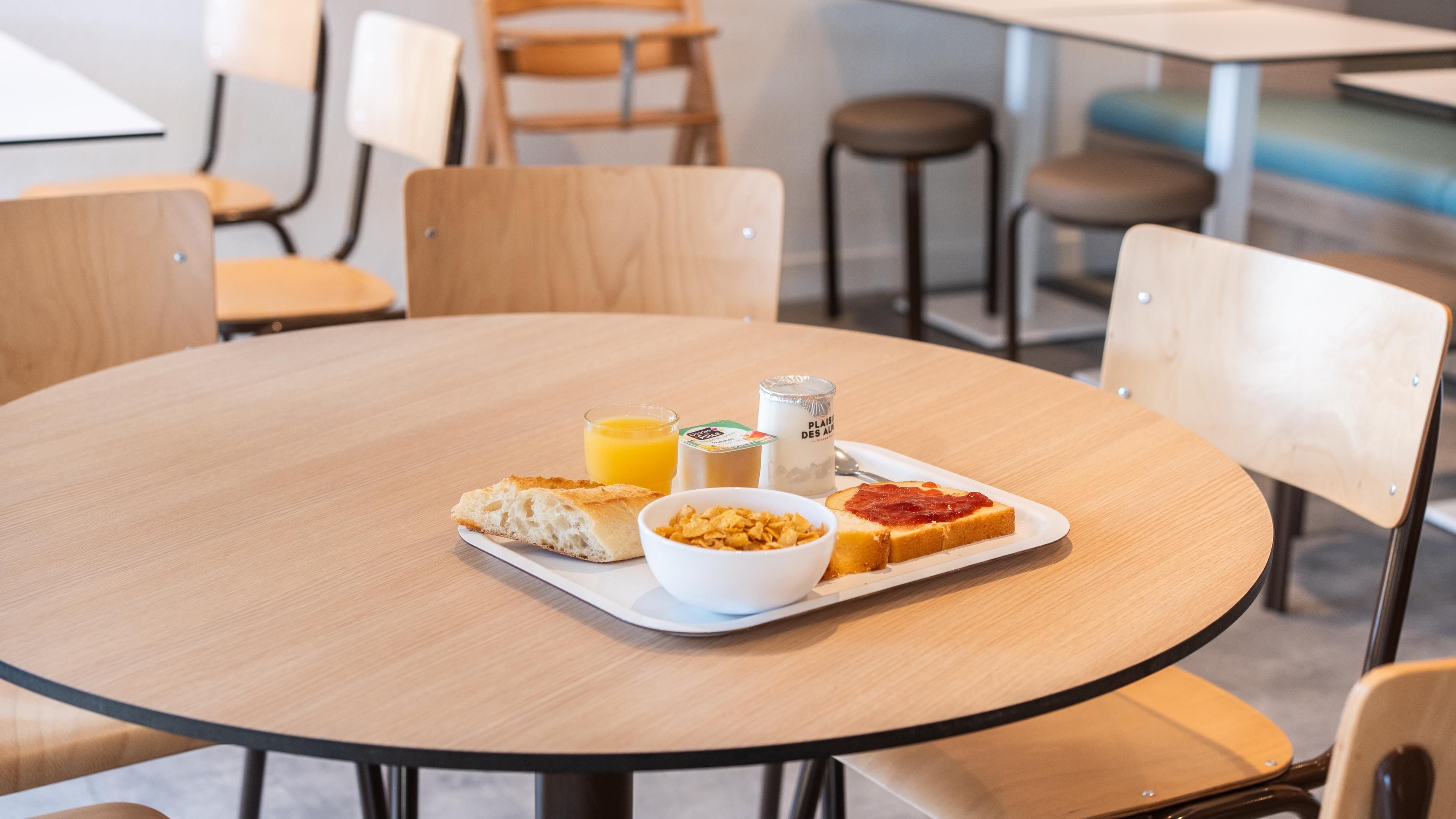 A round wooden table with a breakfast tray containing toast, cereal, a glass of orange juice, and a container of milk.