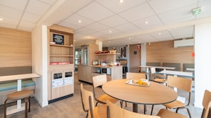 A modern cafeteria with round tables and chairs, a counter with food items, and a wooden wall with a shelf. - Premiere Classe Limoges Nord (Limoges)