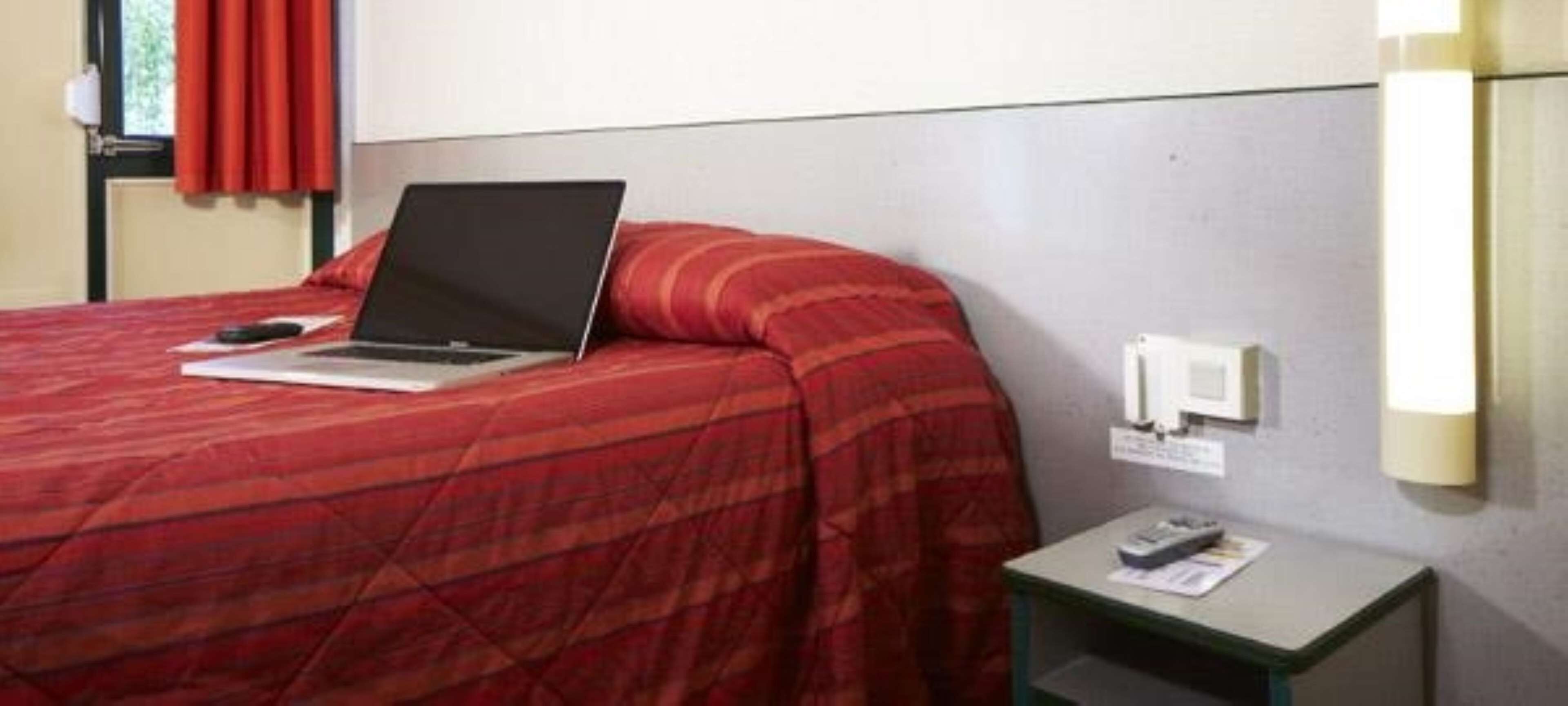 Habitación de hotel con una cama, una laptop sobre la cama, una mesita de noche con una lámpara y una ventana con cortinas.