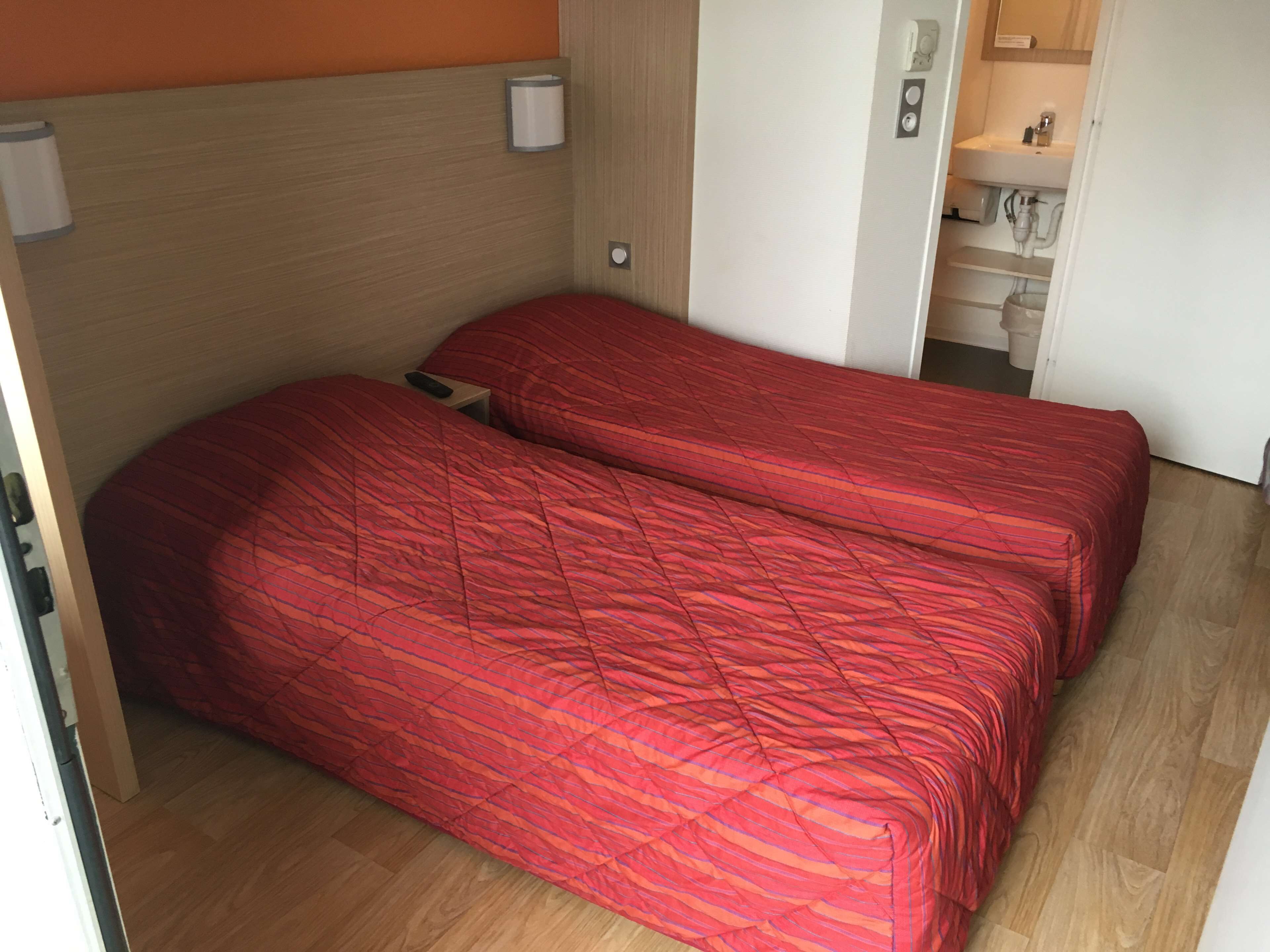 Une chambre avec deux lits, un plancher en bois, une porte blanche et une salle de bain visible, équipée d’un lavabo et d’un miroir.