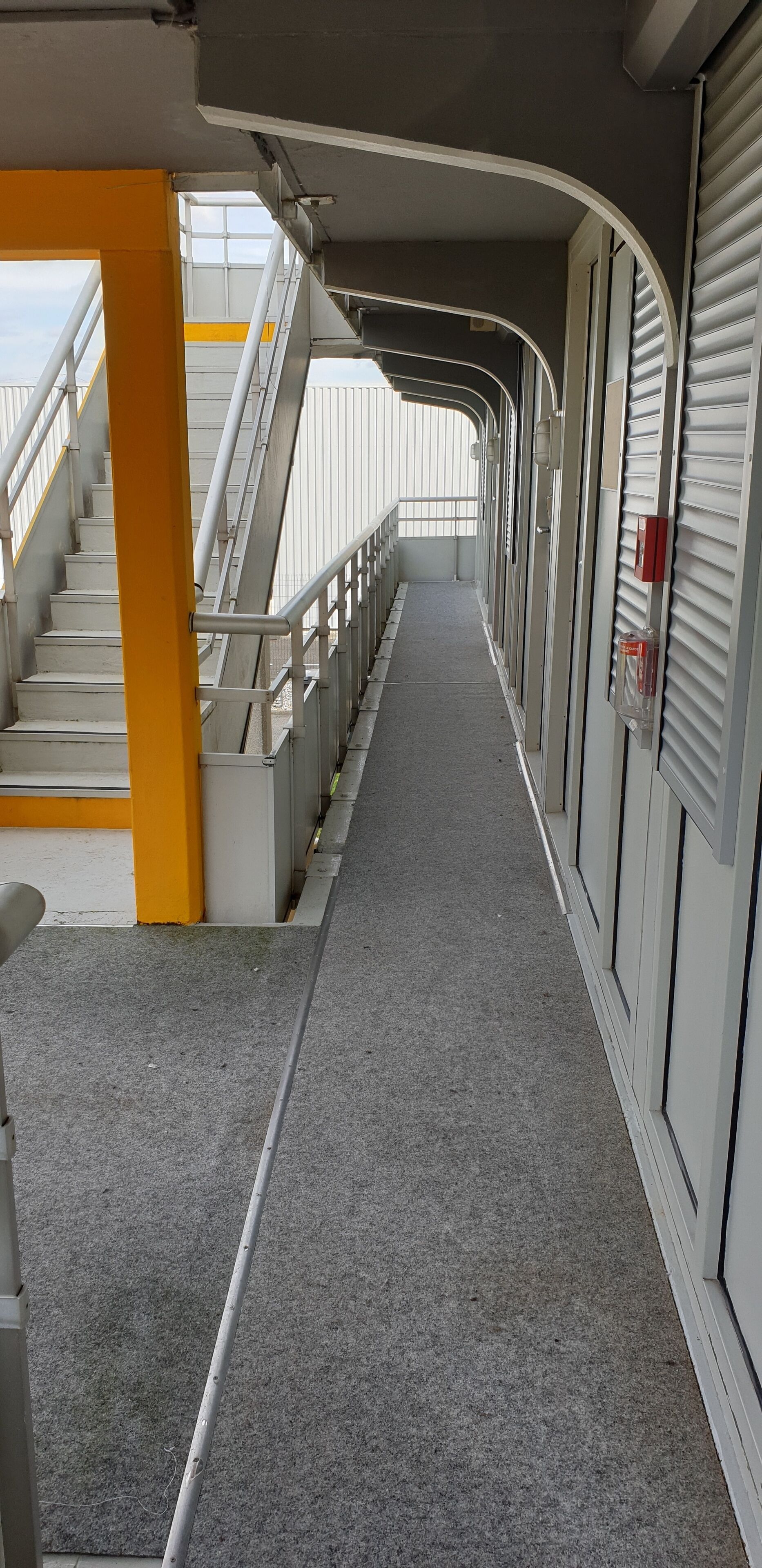 Un pasillo con una escalera a la izquierda, un detector de incendios a la derecha y una vista del cielo a través de una ventana.