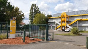 Entrada de hotel con un letrero digital que muestra '52', un edificio amarillo y blanco con una escalera y una cerca verde.