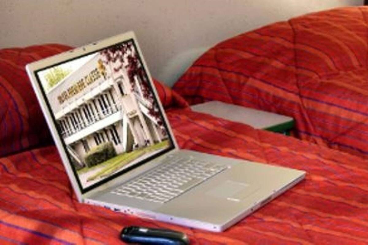 A laptop on a bed with a red blanket, a remote control, and a book.