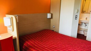 Habitación de hotel pequeña con una cama individual, un escritorio y un baño visible a través de una puerta abierta.