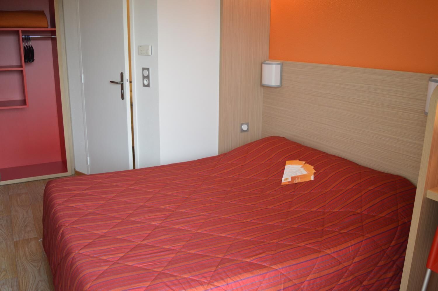 Ein Einzelbett mit roter Bettdecke, eine weiße Tür und ein kleiner Nachttisch.