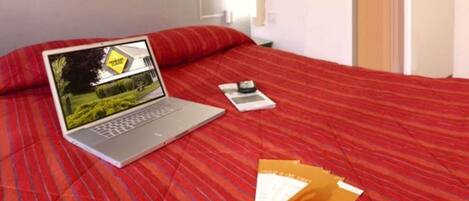Uma cama com um espalhadeira listrada em vermelho, um laptop exibindo uma imagem panorâmica, um livro e alguns papéis sobre a cama.