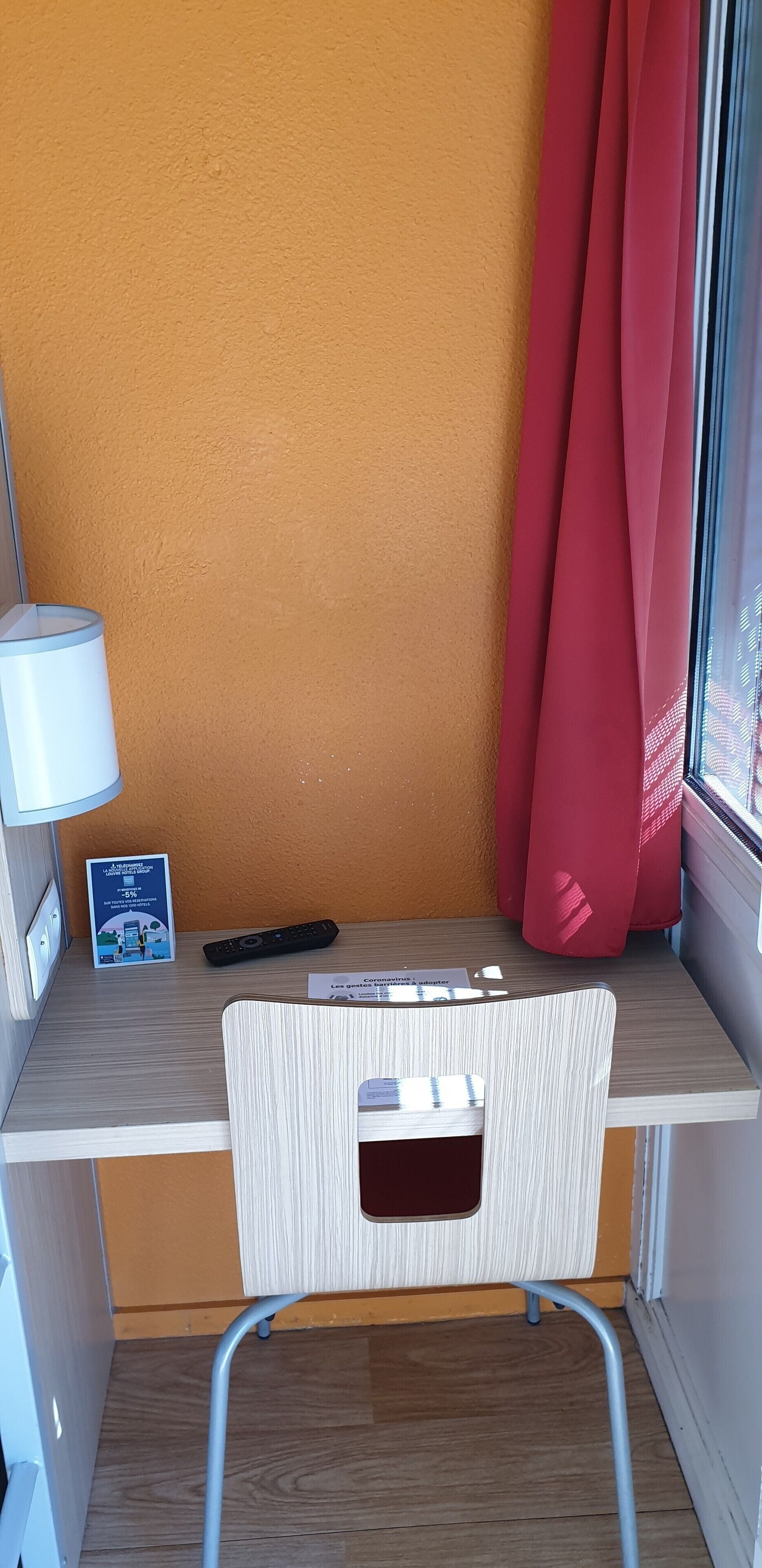 A small, compact workspace with a wooden desk, a white chair, a lamp, a remote control, and a window with red curtains.