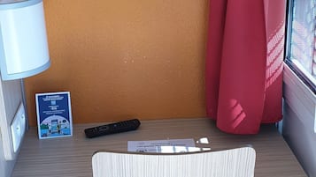 A small, compact workspace with a wooden desk, a white chair, a lamp, a remote control, and a window with red curtains.