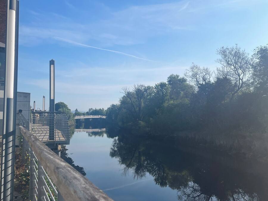 Un río tranquilo con un puente al fondo, una barandilla metálica a lo largo de la orilla y estructuras industriales a la izquierda.
