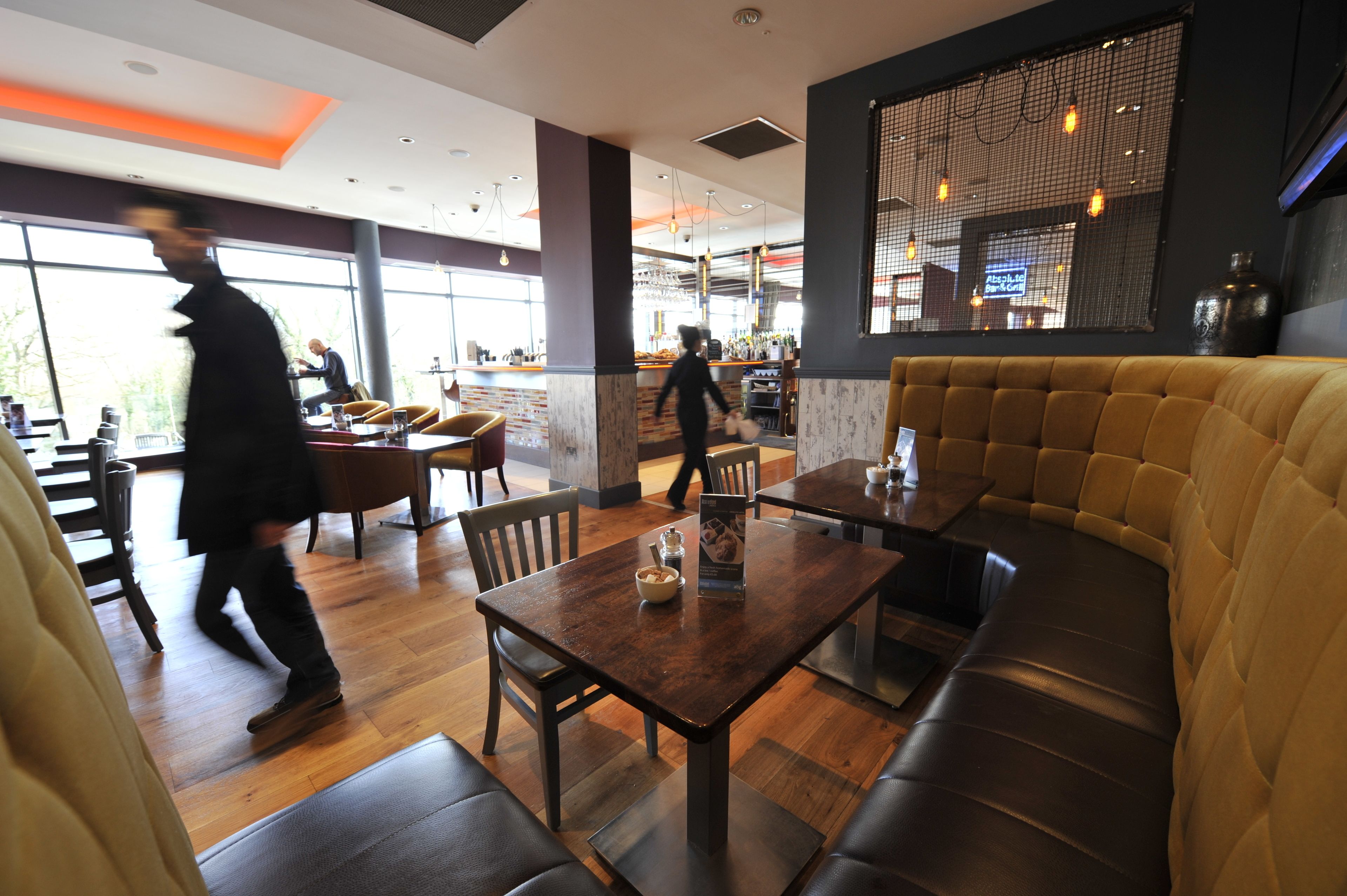 A modern restaurant interior with wooden flooring, a bar area, and a seating arrangement including a booth and tables.