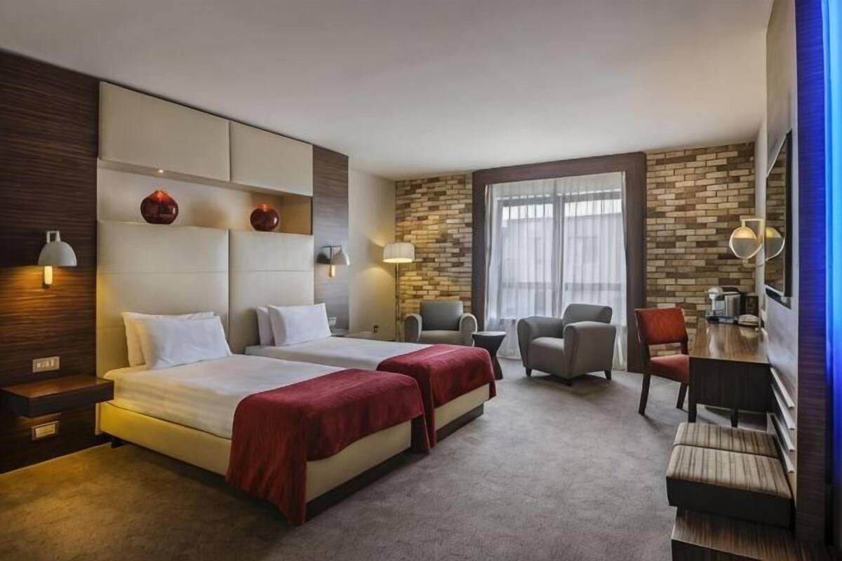 Ett hotellrum med två sängar, ett skrivbord, en stol, en soffa och ett stort fönster.