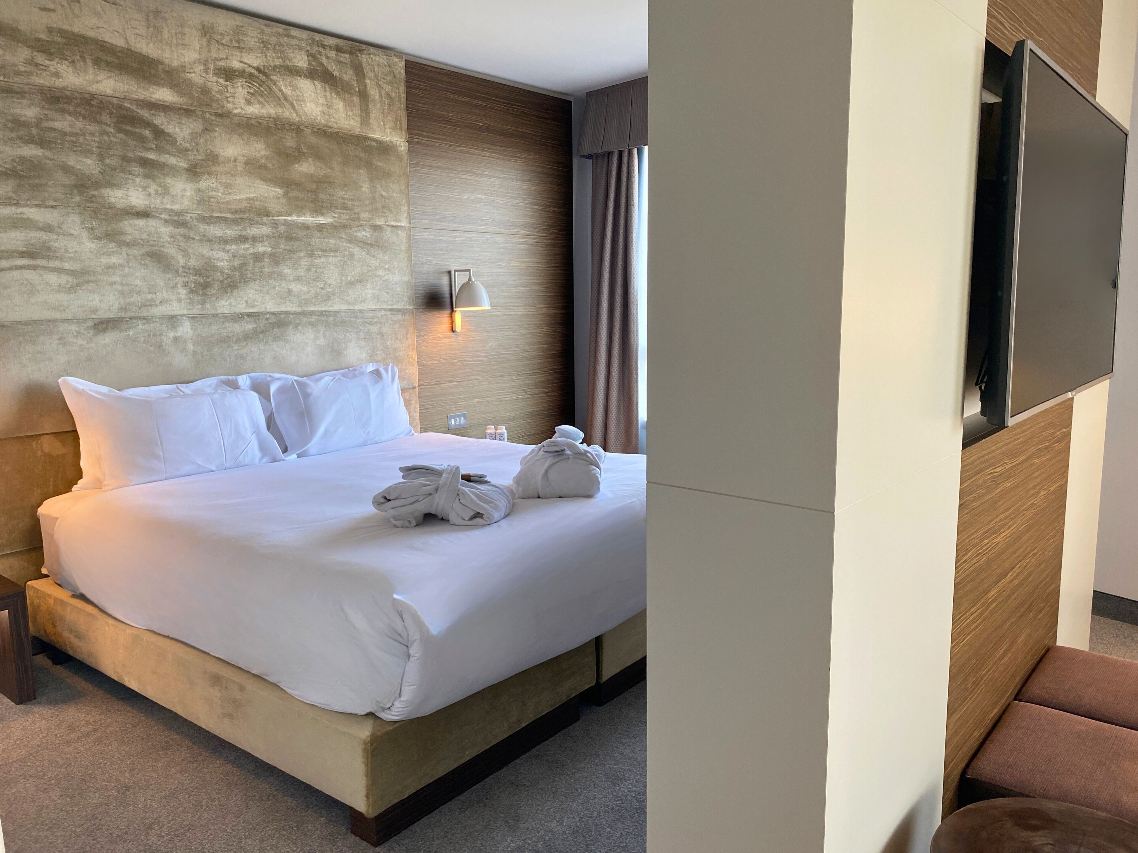 A hotel room with a large bed, a headboard, a wall-mounted TV, and a wooden panel wall.