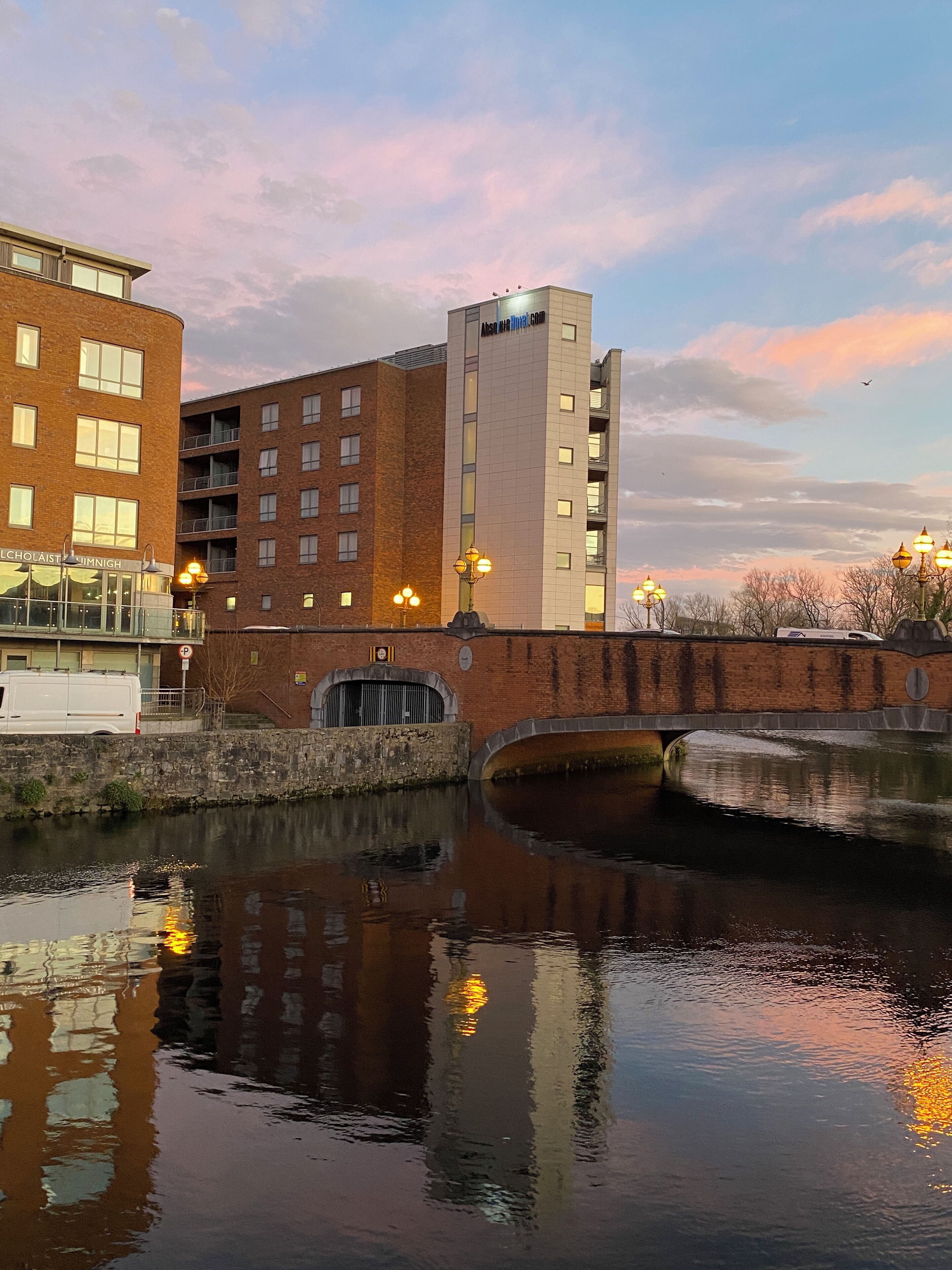 A riverside building with a brick facade, a small bridge, and a reflection in the water.