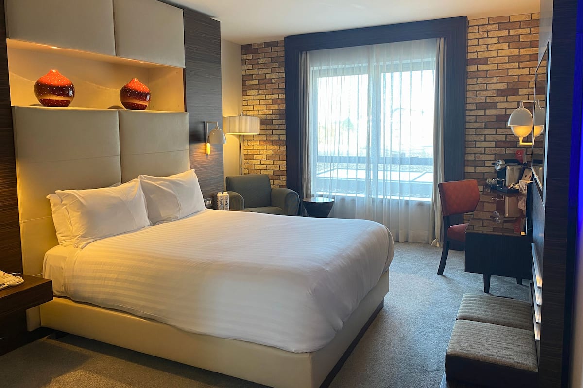 Ett modernt hotellrum med en stor säng, ett skrivbord, en stol och utsikt över omgivningarna genom ett stort fönster.