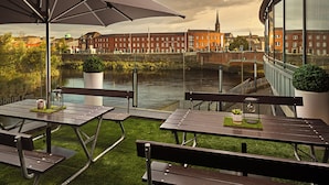 An outdoor patio with picnic tables and umbrellas overlooking a river and historic buildings. - Absolute Hotel Limerick (Limerick)