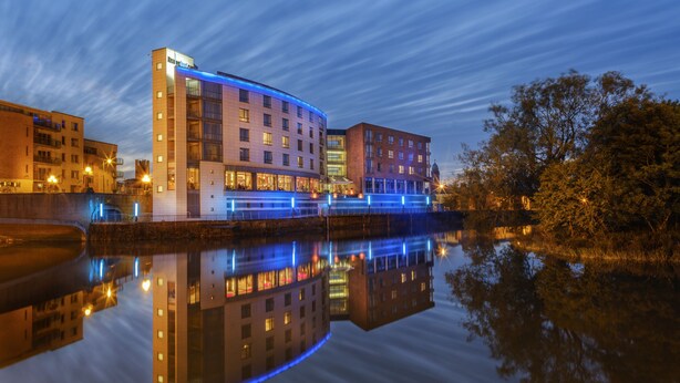 A modern hotel with a reflection in the water, featuring a curved facade and illuminated blue lights. - Absolute Hotel Limerick (Limerick)