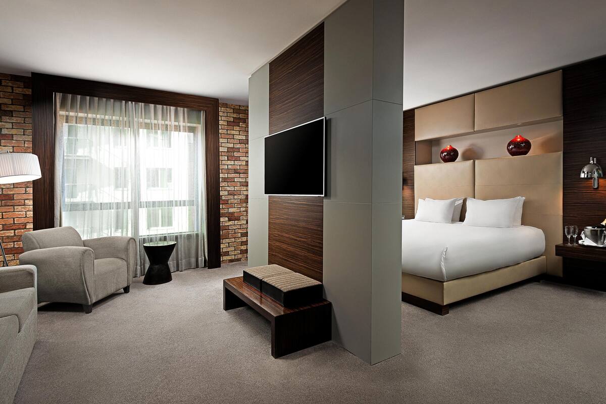 Ett modernt hotellrum med en stor säng, en platt-TV, en vägg med synliga tegelstenar och en sittgrupp med en stol och ett litet bord.