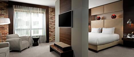 Una habitación de hotel moderna con una cama grande, un televisor de pantalla plana, una pared de ladrillo decorativa y una zona de estar con una silla y una mesita.