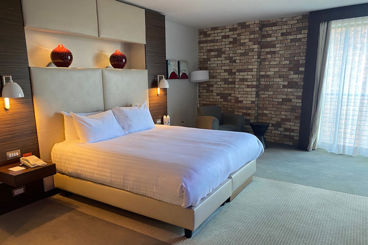 Ett hotellrum med en stor säng, en sänggavel med en lampa, en vägg med ett murstensmönster och ett fönster med gardiner.