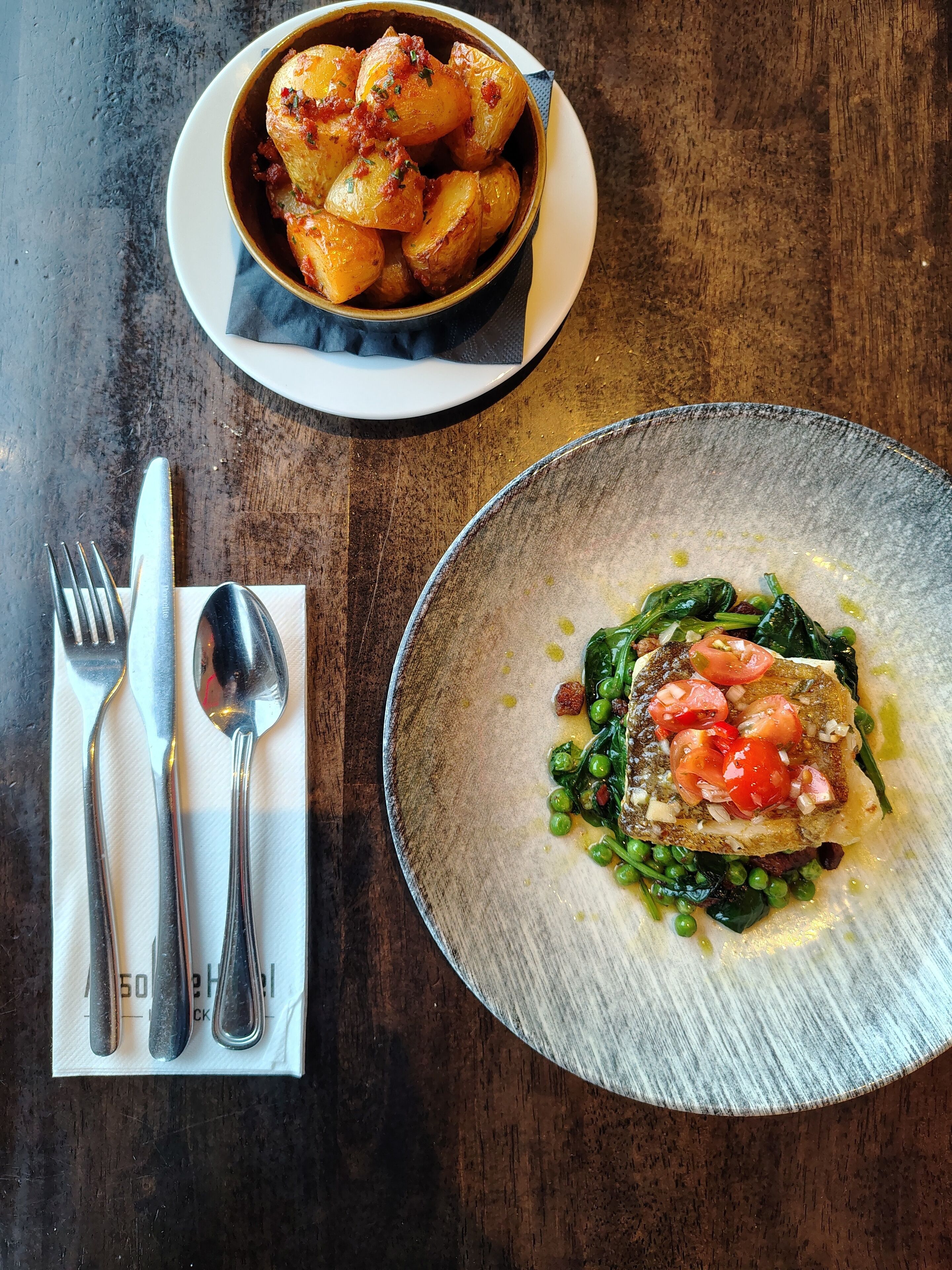 A table with a plate of roasted potatoes and a plate of fish with greens and tomatoes.