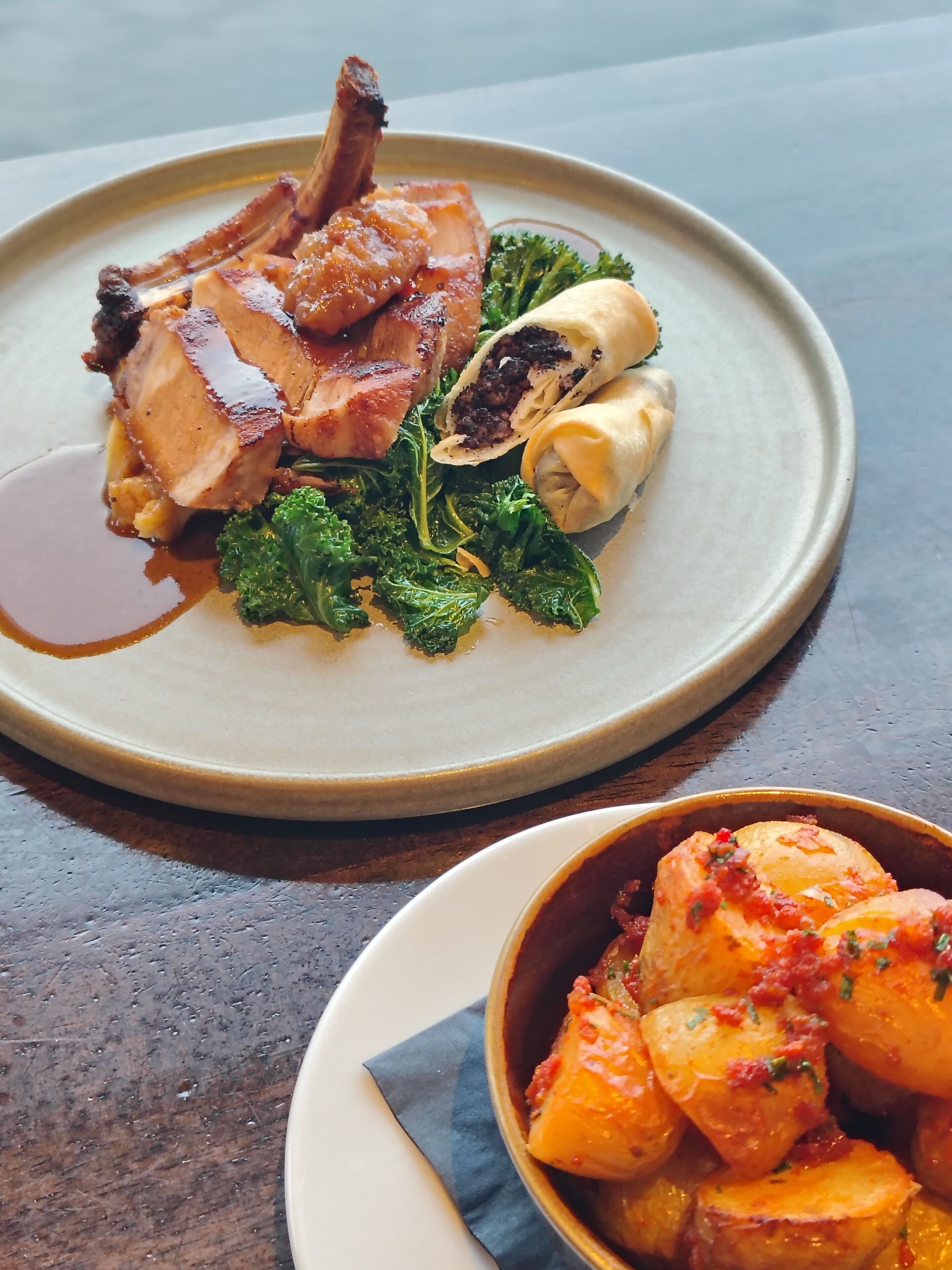 A plate of food with a pork chop, greens, and a rolled item, and a bowl of roasted potatoes.