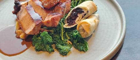 A plate of food with a pork chop, greens, and a rolled item, and a bowl of roasted potatoes.