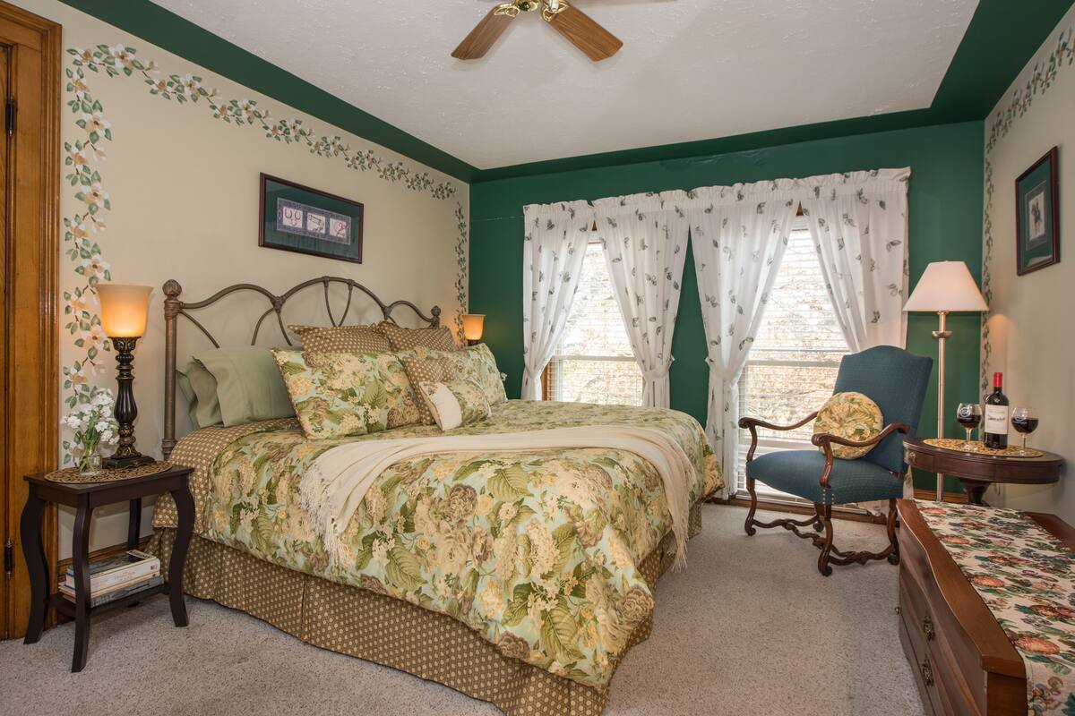 A bedroom with a floral bedspread, a wooden headboard, a side table with a lamp, a chair, and a small table with a wine bottle.