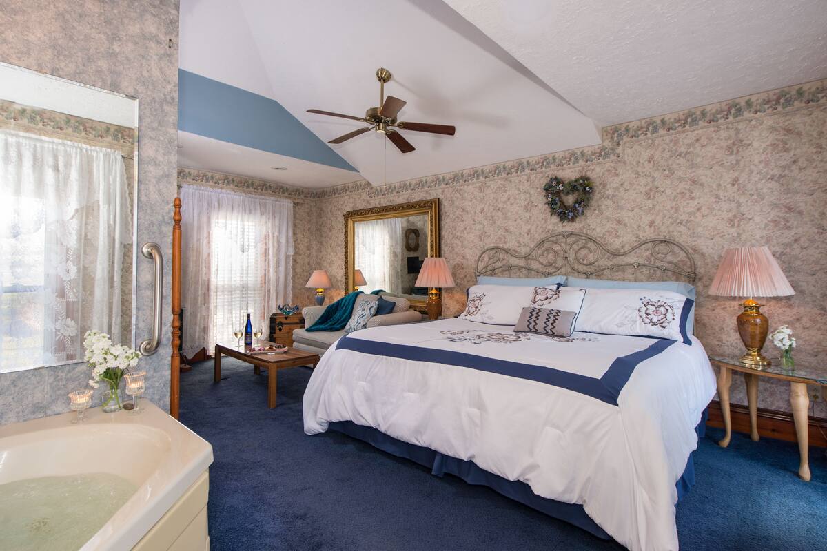 A bedroom with a large bed, a ceiling fan, a mirror, a vanity with a mirror, a bathtub, and a window with curtains.