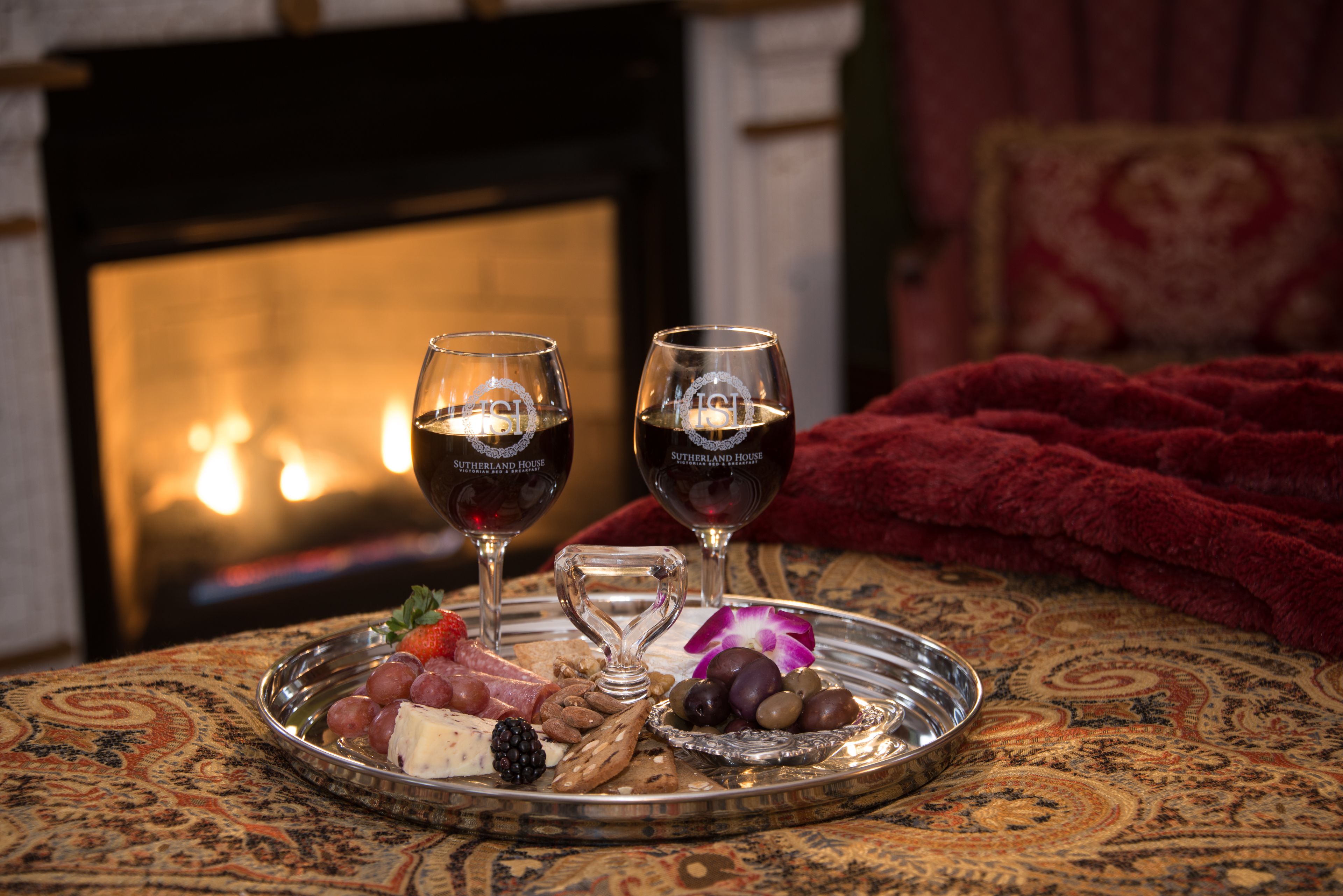 Una bandeja con queso, uvas y aceitunas sobre un mantel estampado, dos copas de vino y una manta roja en una habitación con chimenea.