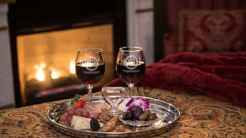 Una bandeja con queso, uvas y aceitunas sobre un mantel estampado, dos copas de vino y una manta roja en una habitación con chimenea.