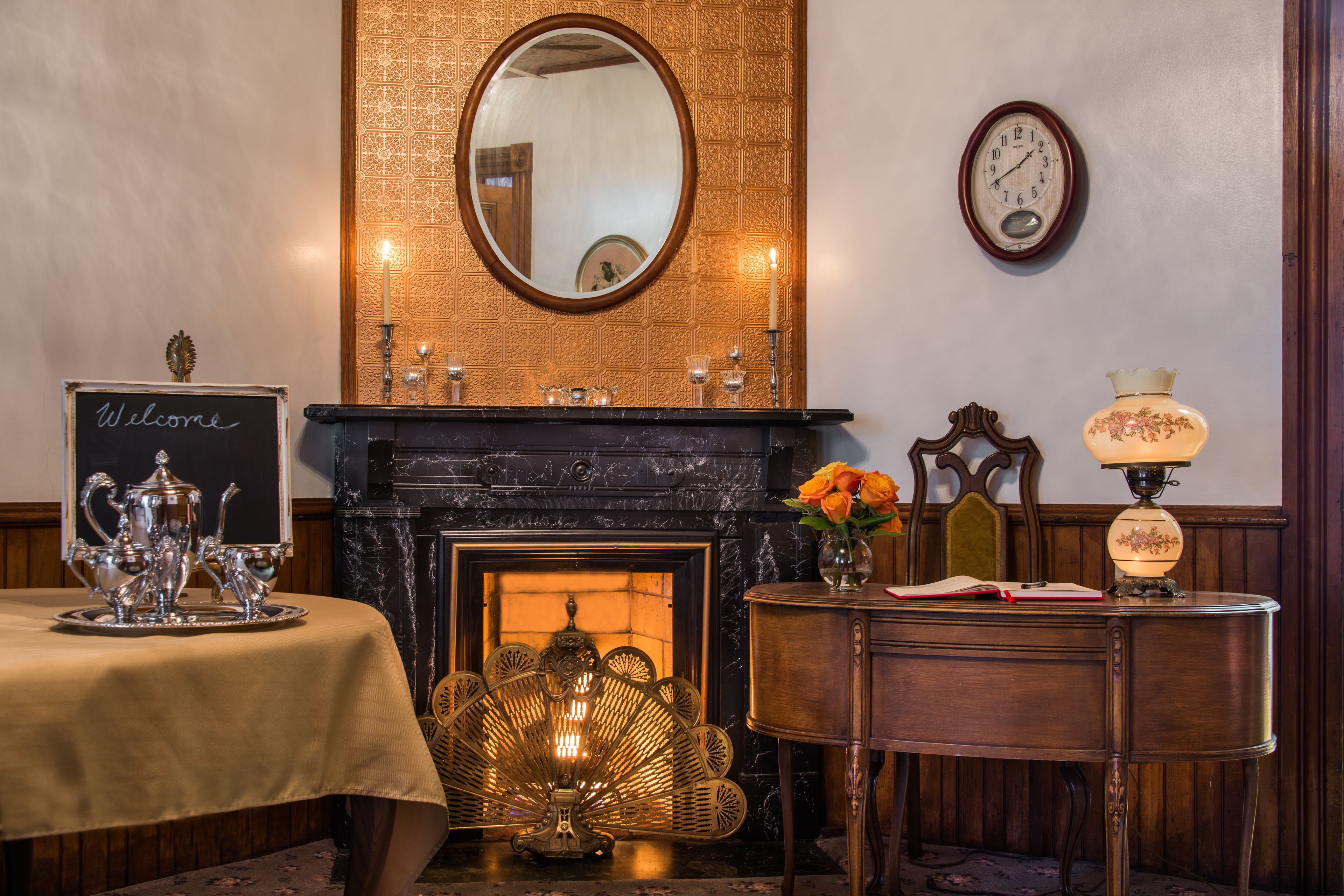 A room with a fireplace, a table with a "Welcome" sign, a mirror, a clock, and a lamp.