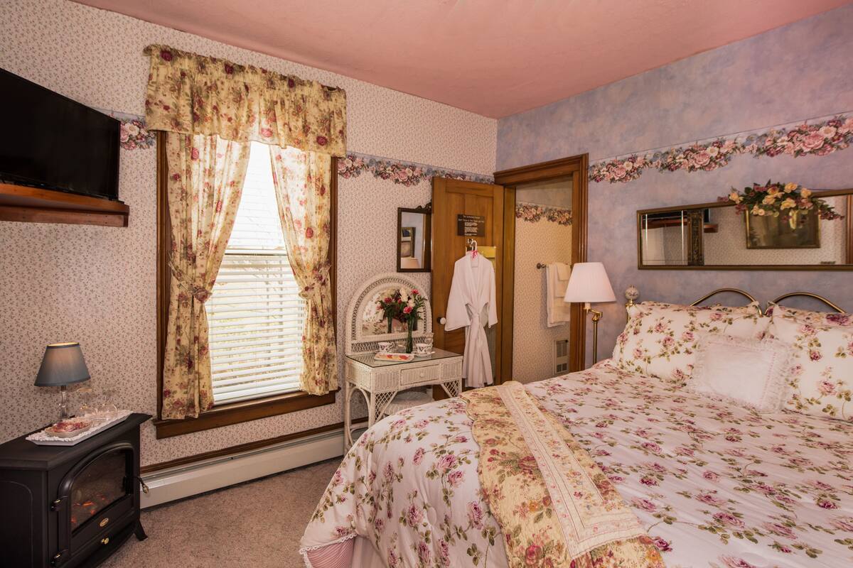 A bedroom with a floral bedspread, a dresser, a vanity with a mirror, and a window with curtains.