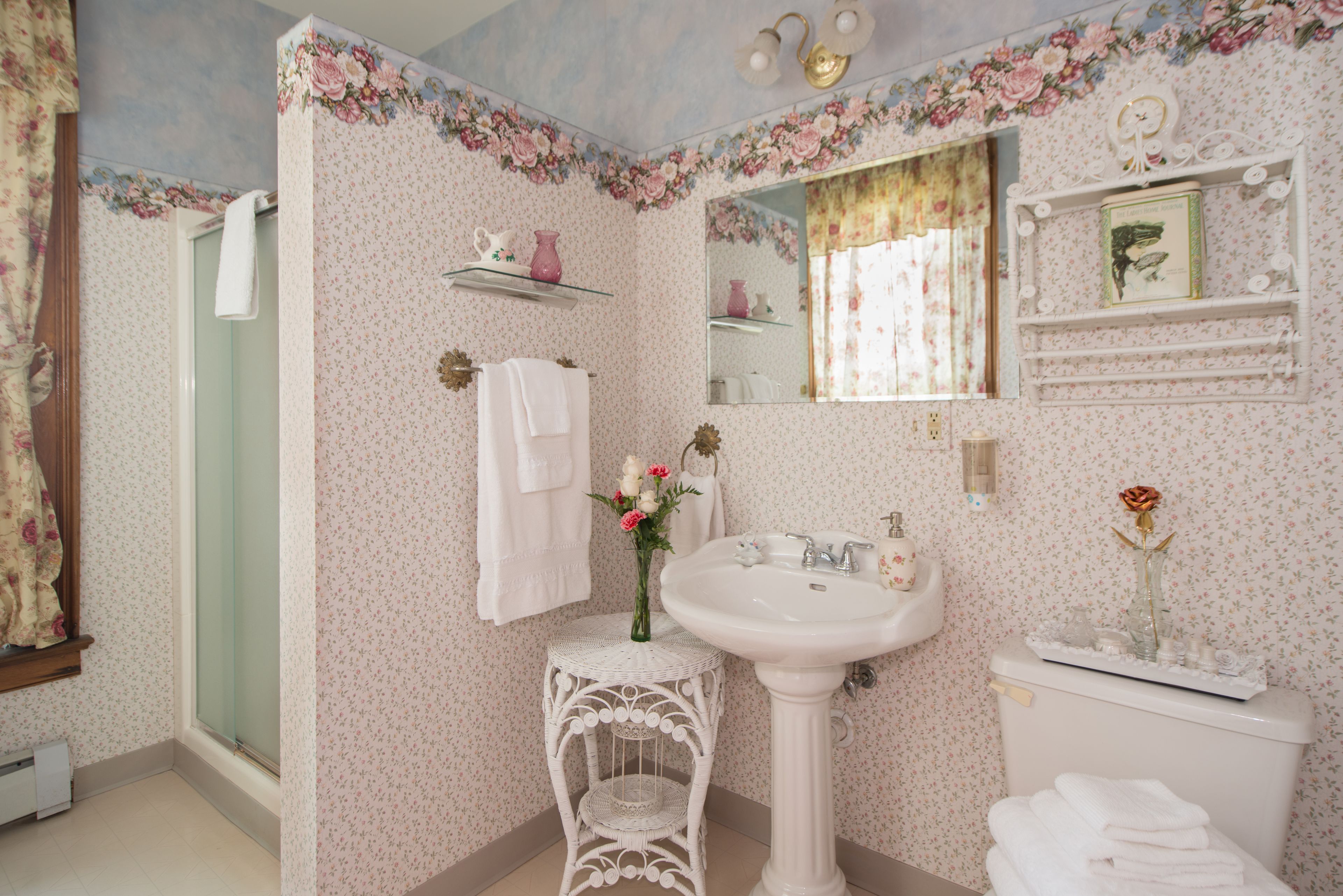 Une salle de bain avec un papier peint à motifs floraux, un lavabo sur colonne, un miroir et des toilettes décorées d’une rose sur le réservoir.