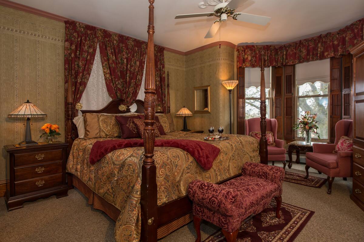 A bedroom with a four-poster bed, a sitting area with two armchairs, a wooden nightstand, and a mirror.