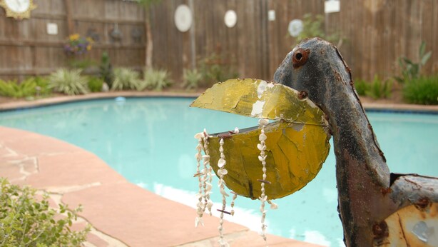 A rustic metal sculpture with a leaf and beads, positioned near a swimming pool with a wooden fence and plants in the background.