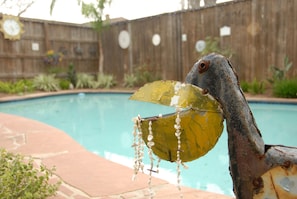 A rustic metal sculpture with a leaf and beads, positioned near a swimming pool with a wooden fence and plants in the background.