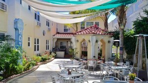 Une terrasse extérieure avec plusieurs tables et chaises, un espace de repos couvert et un bâtiment sur lequel on peut lire l’inscription « HÔTEL ».