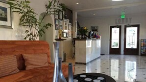 A lobby with a sofa, a reception desk, framed pictures, and a large plant. - El Primero Hotel (Chula Vista)